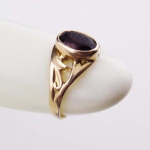 May include: A gold ring featuring an oval-shaped, deep purple gemstone set in a bezel. The band displays an openwork, swirling metal design that extends from the setting down the sides of the ring, showcasing a polished metallic finish.