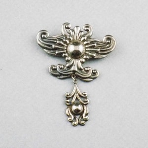 May include: Silver-toned brooch with an ornate, symmetrical design. The brooch features a central circular element with radiating stylized floral or foliate motifs. A smaller, decorative pendant hangs below.