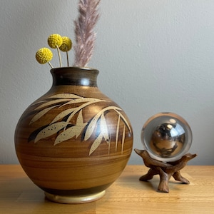 Może przedstawiać: Brązowy ceramiczny wazon z wzorem liści, w którym dominuje jasnobrązowy i biały wzór. Wazon jest wypełniony suszonymi żółtymi kwiatami i gałązką suszonej trawy. Wazon stoi na drewnianej powierzchni.