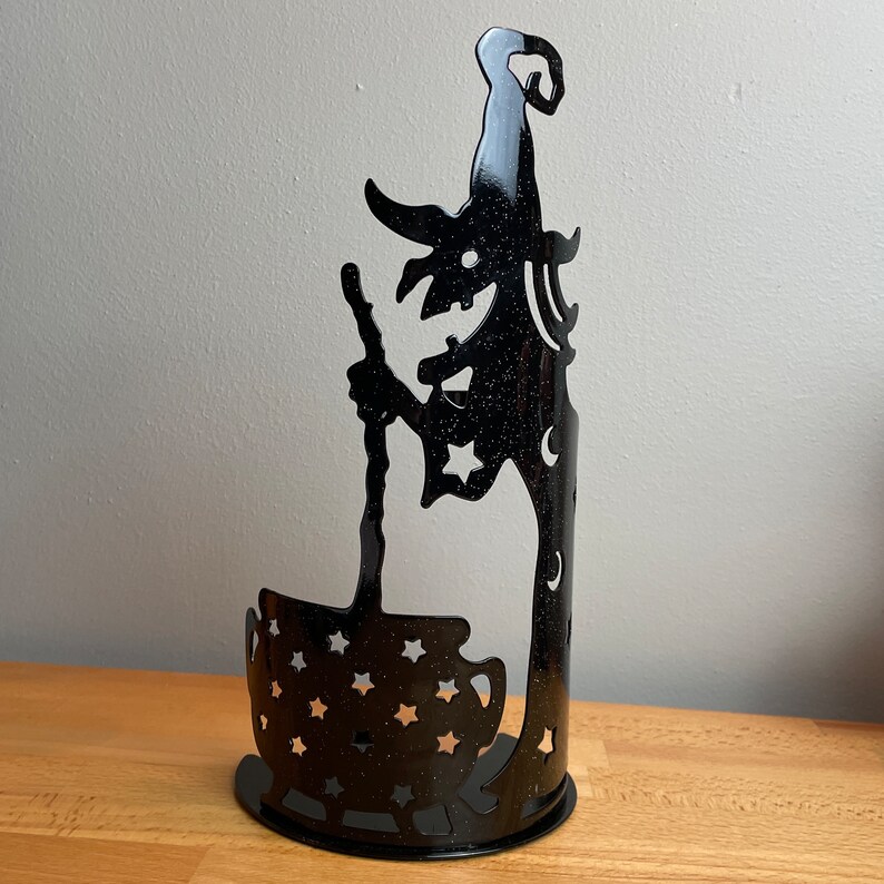 May include: Black metal Halloween candle holder shaped like a witch holding a cauldron. The witch is silhouetted against a starry sky and the cauldron is decorated with stars.