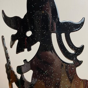 May include: Black metal silhouette of a witch holding a broom. The silhouette is covered in black glitter.