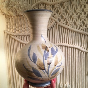 May include: A beige ceramic vase with a bulbous body and a slender neck. The vase features a blue rim and base, with a painted floral design in blue and tan. The vase is approximately 10 inches tall.