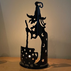 May include: Black metal candle holder shaped like a witch holding a cauldron. The witch has a pointed hat and stars and moons on her dress. The cauldron has stars on it.