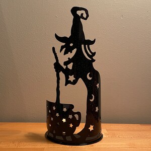 May include: Black metal candle holder shaped like a witch holding a cauldron. The witch has a pointed hat and stars and moons on her dress. The cauldron has stars on it.