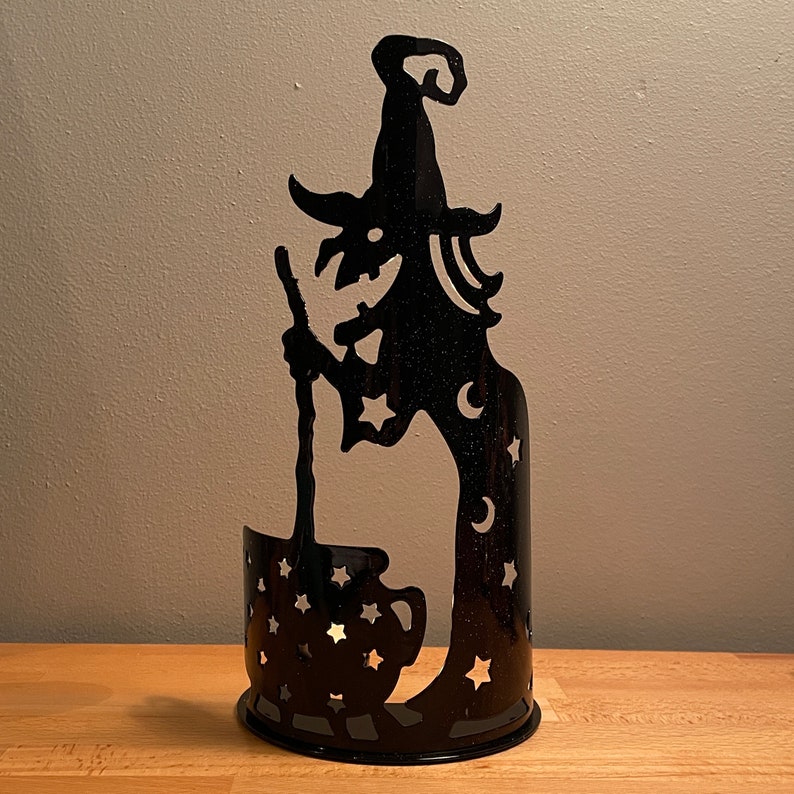 May include: Black metal candle holder shaped like a witch holding a cauldron. The witch has a pointed hat and stars and moons on her dress. The cauldron has stars on it.