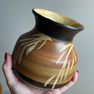 Może przedstawiać: Ceramiczny wazon w odcieniach brązu i beżu, z czarną obwódką i złotym wnętrzem. Wazon ma unikalny kształt i jest ozdobiony jasnobeżowym wzorem liści. Wazon jest trzymany przez osobę.