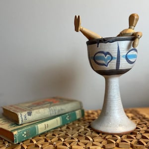 May include: A decorative ceramic chalice with a white base, black rim, and blue heart and stripe designs. A wooden artist's mannequin is positioned inside the chalice. Two vintage books are in the foreground.