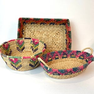May include: Three woven baskets with colorful trim. The baskets are made of natural materials and have a rustic, handmade look. The largest basket is rectangular, while the other two are round and oval.