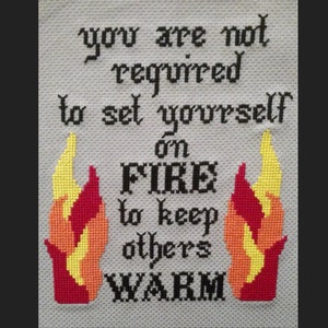 May include: Cross-stitch embroidery with the text "you are not required to set yourself on fire to keep others warm" and two red and yellow flames on either side.