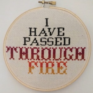 Pode incluir: Um aro de bordado branco com a frase "I have passed through fire" bordada em fio preto e vermelho. A palavra "fire" está em um degradado de vermelho, laranja e amarelo.
