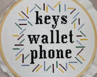 May include: A white embroidery hoop with a wooden frame. The embroidery features a white background with black text that reads "keys wallet phone" and a colorful border of stitched lines.