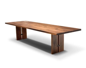 May include: A long, rectangular wooden table with a natural wood finish. The table has a simple design with two sets of legs that are attached to the underside of the table top.