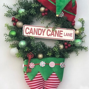 May include: A Christmas wreath with a green base and red and gold ornaments. A sign that reads "CANDY CANE LANE" is attached to the wreath. An elf's legs and feet are hanging from the bottom of the wreath.