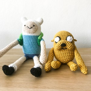 Puede incluir: Dos juguetes de peluche tejidos a crochet, uno azul y blanco con una bufanda verde, y otro amarillo con ojos blancos.