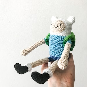 Op de afbeelding: Gehaakt pluche speelgoed van Finn de Mens uit de animatieserie Adventure Time. Finn draagt een blauw shirt en een groene rugzak. Hij heeft een wit gezicht met zwarte ogen en een glimlach.