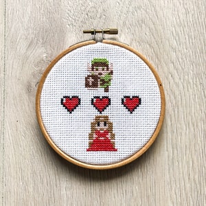 Op de afbeelding: Borduurwerk in een hoepel met een gepixelde afbeelding van een videogamepersonage, Link, uit de Legend of Zelda-serie, met een hart boven zijn hoofd. Daaronder is een gepixelde afbeelding van een vrouwelijk personage met een hart boven haar hoofd. De hoepel is gemaakt van hout en heeft een bruine afwerking.