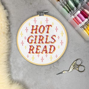 Op de afbeelding: Een ronde houten borduurring met witte Aida-stof waarop de tekst HOT GIRLS READ in kruissteek is geborduurd in een verloop van roze, oranje en geel. Het ontwerp is versierd met kleine roze en gele kruissteek sterretjes.