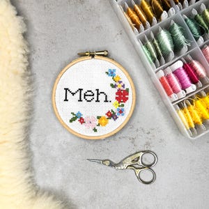 Op de afbeelding: Een ronde houten borduurring met witte Aida-stof en een kruissteekontwerp. De tekst "Meh." is in het zwart geborduurd, omringd door een kleurrijke bloemenkrans van rood, blauw, geel en groen garen. Er ligt een metalen schaar bij.