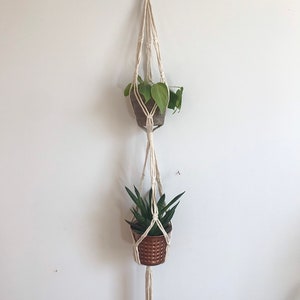 May include: A white macrame plant hanger with two hanging pots. The top pot is brown and holds a green plant with heart-shaped leaves. The bottom pot is copper and holds a green succulent.