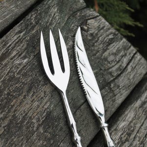 May include: Close-up of a silver fork and knife set. The fork has three tines, and the knife has a serrated edge. The handles have a decorative design. The silverware is placed on a weathered wooden surface.