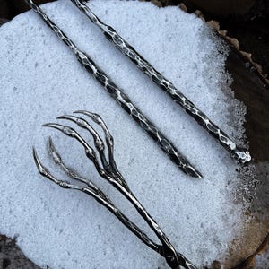 May include: Hand-forged metal fire tools, including tongs and pokers, rest on a snowy surface. The tools have a textured, hammered appearance and are designed for use with a fireplace or outdoor fire pit. The tongs feature a claw-like design.