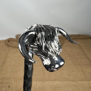 May include: A silver sculpture of a Highland cow's head. The sculpture has detailed fur and curved horns, mounted on a textured, dark metal pole. The cow's head is the focal point, with a shiny, reflective surface.