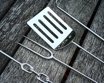 May include: A set of four stainless steel grilling tools, including a spatula, a fork, a chain, and a long, thin tool. The tools are laid out on a wooden surface.