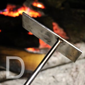 May include: A stainless steel fire pit tool with a rectangular head and a long handle. The tool is designed for use in a fire pit, with a blurred background of a burning fire. The letter "D" is visible in the lower left corner.