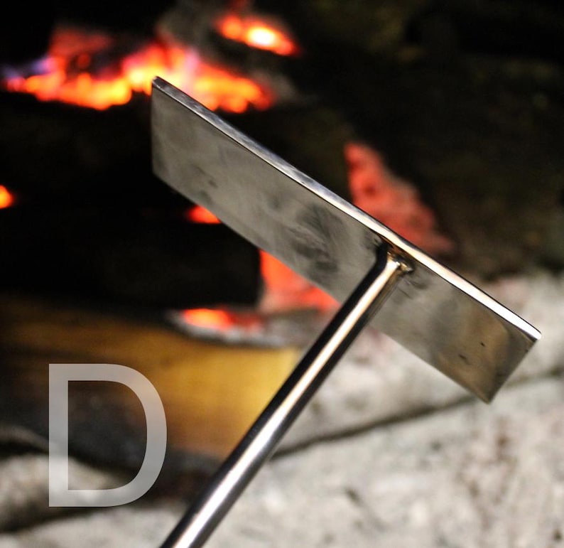 May include: A stainless steel fire pit tool with a rectangular head and a long handle. The tool is designed for use in a fire pit, with a blurred background of a burning fire. The letter "D" is visible in the lower left corner.