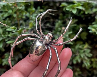 May include: A detailed, silver-colored spider sculpture with articulated legs and a rounded body. The spider is held in a hand against a backdrop of green foliage and a spider web. The sculpture has a reflective surface.