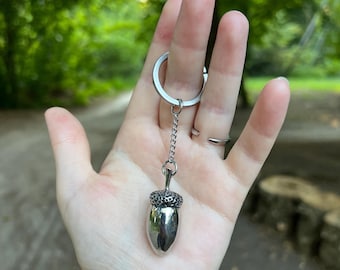 May include: A silver-toned metal keychain featuring a realistic acorn charm. The acorn has a textured, dark cap and a smooth, polished nut body. It is attached to a standard circular split ring by a short, delicate silver-colored metal chain.