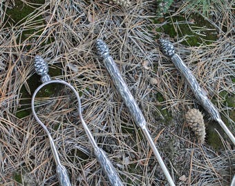 May include: Silver-toned fireplace tools set, including a poker, shovel, and tongs, with pinecone-shaped handles. The tools are arranged on a bed of pine needles and small pine cones.