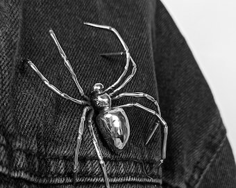 May include: A silver spider brooch with a detailed design. The brooch is pinned to a denim jacket.