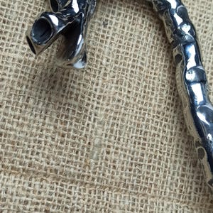 May include: A detailed, silver-colored dragon-shaped handle, possibly for a cane or walking stick, is displayed against a textured, tan background. The dragon's head is prominent, with open mouth and detailed features.