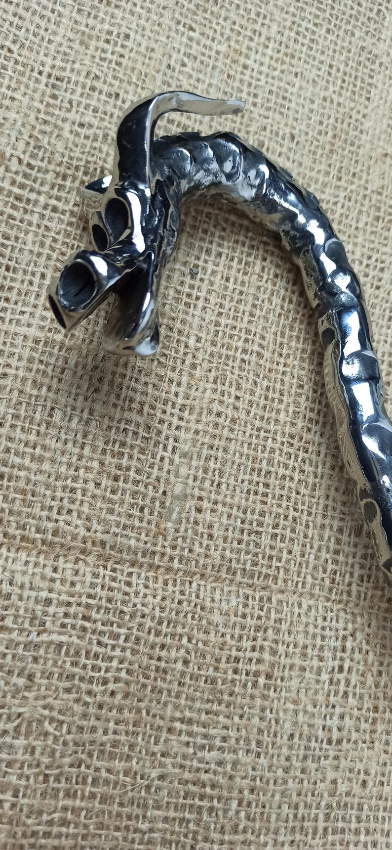 May include: A detailed, silver-colored dragon-shaped handle, possibly for a cane or walking stick, is displayed against a textured, tan background. The dragon's head is prominent, with open mouth and detailed features.