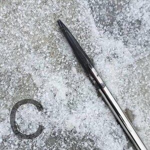 May include: Close-up of a knitting needle with a dark, pointed tip and a shiny, silver shaft. The needle is angled across a textured, snow-covered surface. A dark gray letter "C" is in the lower left corner.