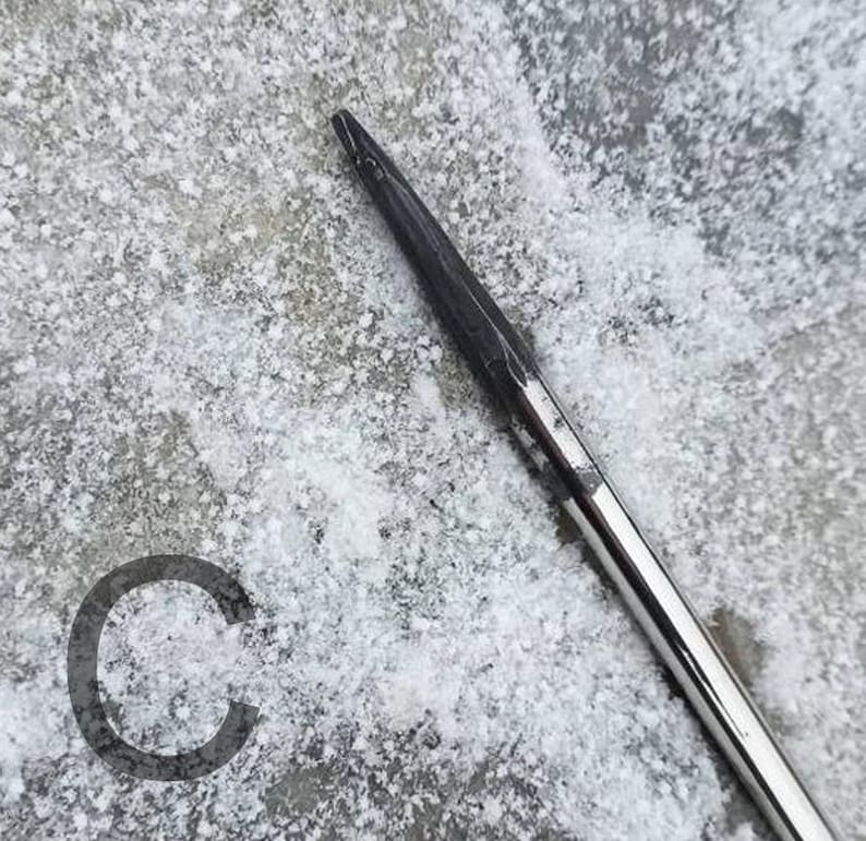 May include: Close-up of a knitting needle with a dark, pointed tip and a shiny, silver shaft. The needle is angled across a textured, snow-covered surface. A dark gray letter "C" is in the lower left corner.