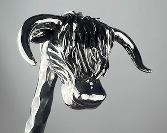 May include: A polished silver sculpture of a Highland cow's head. The sculpture features detailed fur and curved horns. The cow's head is angled slightly downward, showcasing the intricate craftsmanship of the metalwork.
