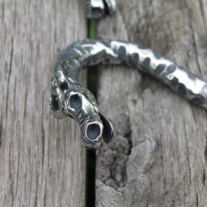 May include: Close-up of a silver-toned metal dragon-shaped object. The dragon's head is detailed with open eye sockets and a curved, textured body. The object is set against a weathered wooden surface.