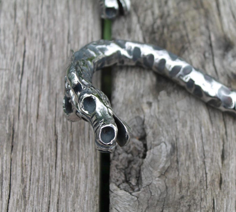 May include: Close-up of a silver-toned metal dragon-shaped object. The dragon's head is detailed with open eye sockets and a curved, textured body. The object is set against a weathered wooden surface.