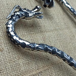 May include: Two silver-colored, dragon-shaped bottle openers. The openers have a textured, hammered metal appearance. The dragon heads are detailed, with open mouths and visible features. The openers are on a textured, tan fabric.