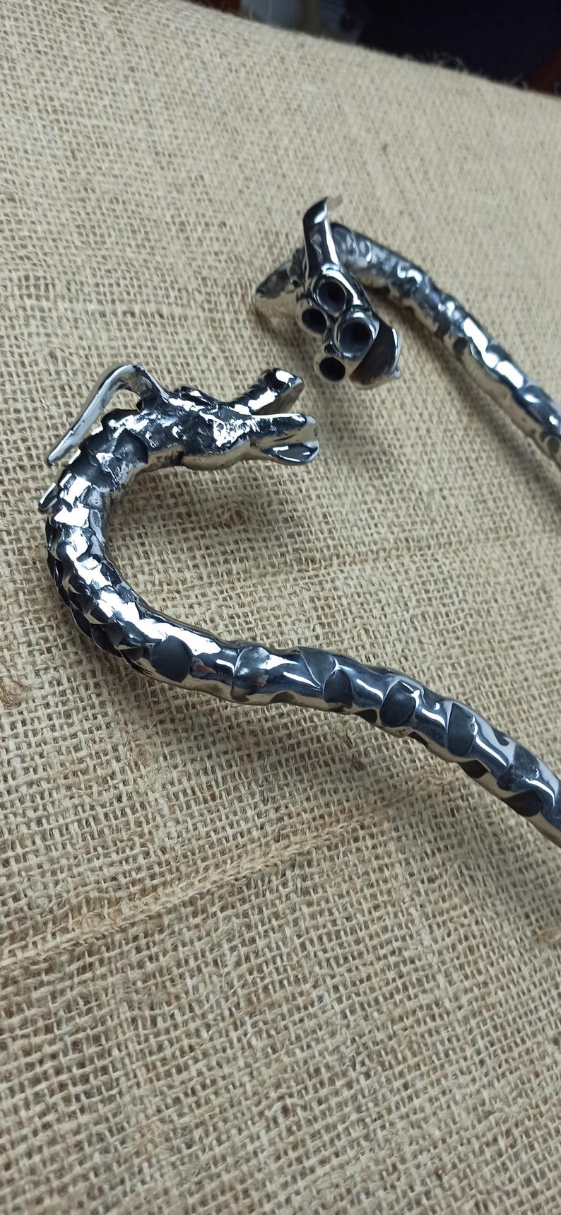 May include: Two silver-colored, dragon-shaped bottle openers. The openers have a textured, hammered metal appearance. The dragon heads are detailed, with open mouths and visible features. The openers are on a textured, tan fabric.