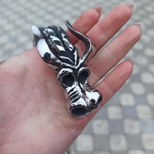 May include: A silver-colored ring with a stylized dragon head design. The ring features a detailed dragon head with horns, eyes, and a snout. The ring is held in a hand, showcasing its intricate craftsmanship and metallic sheen.