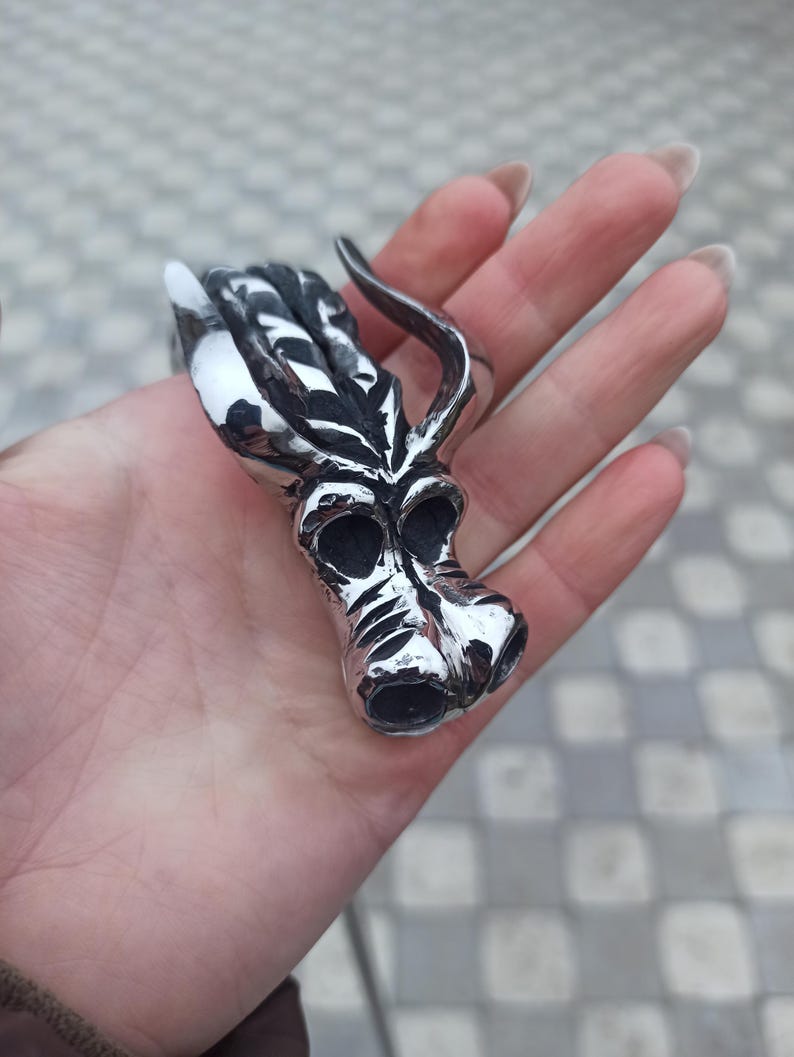 May include: A silver-colored ring with a stylized dragon head design. The ring features a detailed dragon head with horns, eyes, and a snout. The ring is held in a hand, showcasing its intricate craftsmanship and metallic sheen.