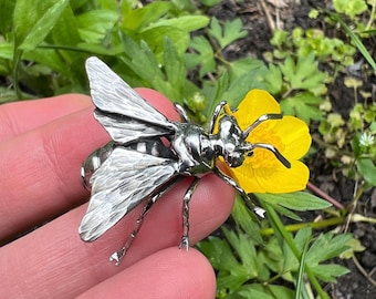 May include: A detailed silver-colored bee brooch with textured wings and body, resting on a bright yellow flower. The brooch is intricately designed, showcasing the insect's features. The background features green foliage.