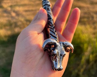 May include: A silver-colored wand with a ram's head design. The wand has a braided handle and a pointed tip. The ram's head is detailed with horns and facial features. The wand is held in a hand.