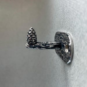 May include: A silver-colored wall hook featuring a detailed pinecone design. The hook has a textured, branch-like arm extending from a rectangular mounting plate. The pinecone is at the end of the arm.