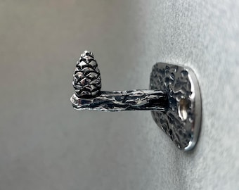 May include: A silver-colored wall hook featuring a detailed pinecone design. The hook has a textured, branch-like arm extending from a rectangular mounting plate. The pinecone is at the end of the arm.