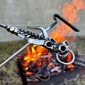 May include: A silver metal dragon head skewer with a ring attached to its mouth. The skewer is over a fire pit with flames. The dragon head has detailed features and is made of shiny metal.