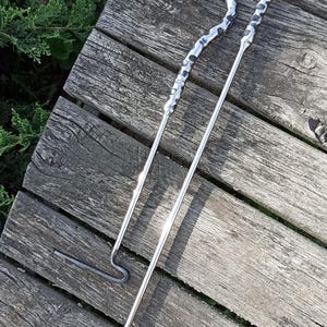 May include: Two metal fire pit tools with dragon head designs. One is a poker, and the other is a hook. The tools are silver and rest on weathered wooden planks.